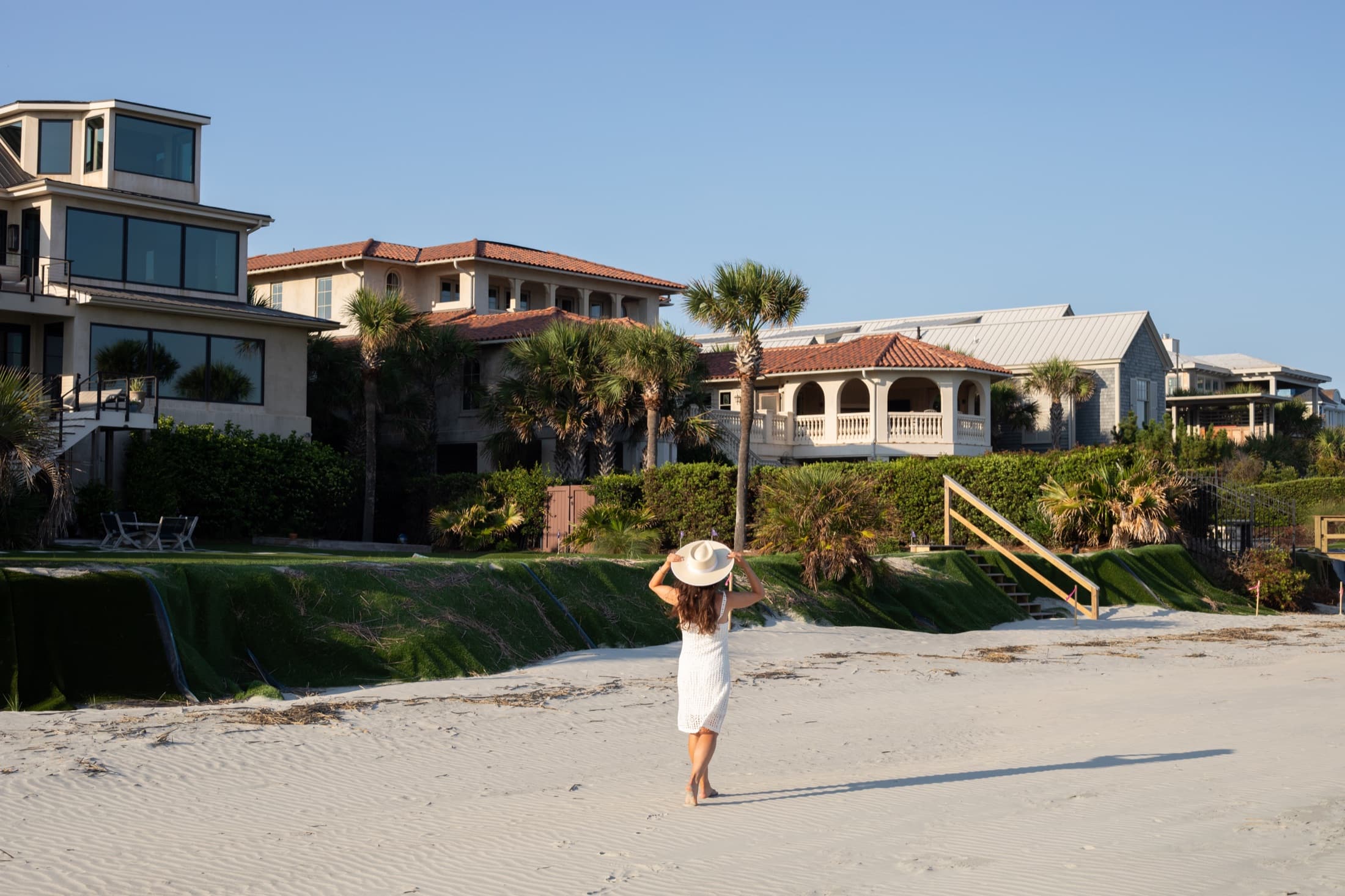 Woman in a white dress and wide-brim hat walking along an empty beach at golden hour, the shoreline stretching ahead of her