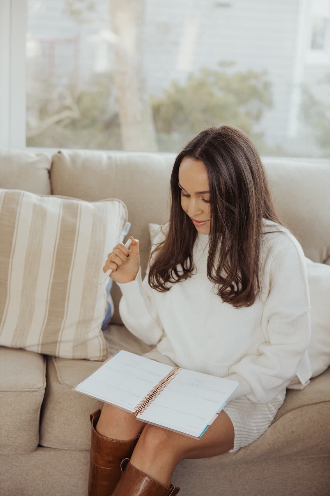 Woman in a cream sweater sitting on a sofa, pen in hand, working through a guided workbook in soft window light